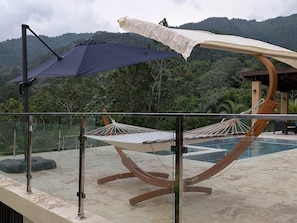 A hammock under a large umbrella, a pool, and a mountainous backdrop.