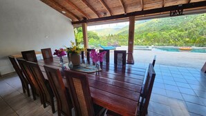 A wooden dining table with chairs, a vase with flowers, and a view of a pool and mountains.