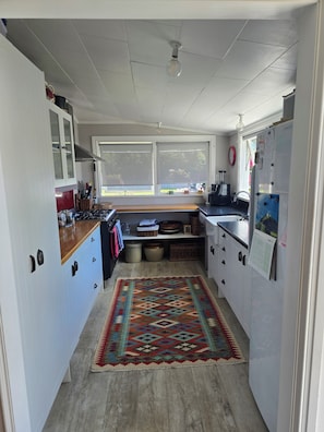 A compact kitchen with white cabinets, a wooden countertop, a patterned rug, and a window with a view of the outdoors. - Willowstream Cottage - Te Awanga, Hawkes Bay NZ (Te Awanga)
