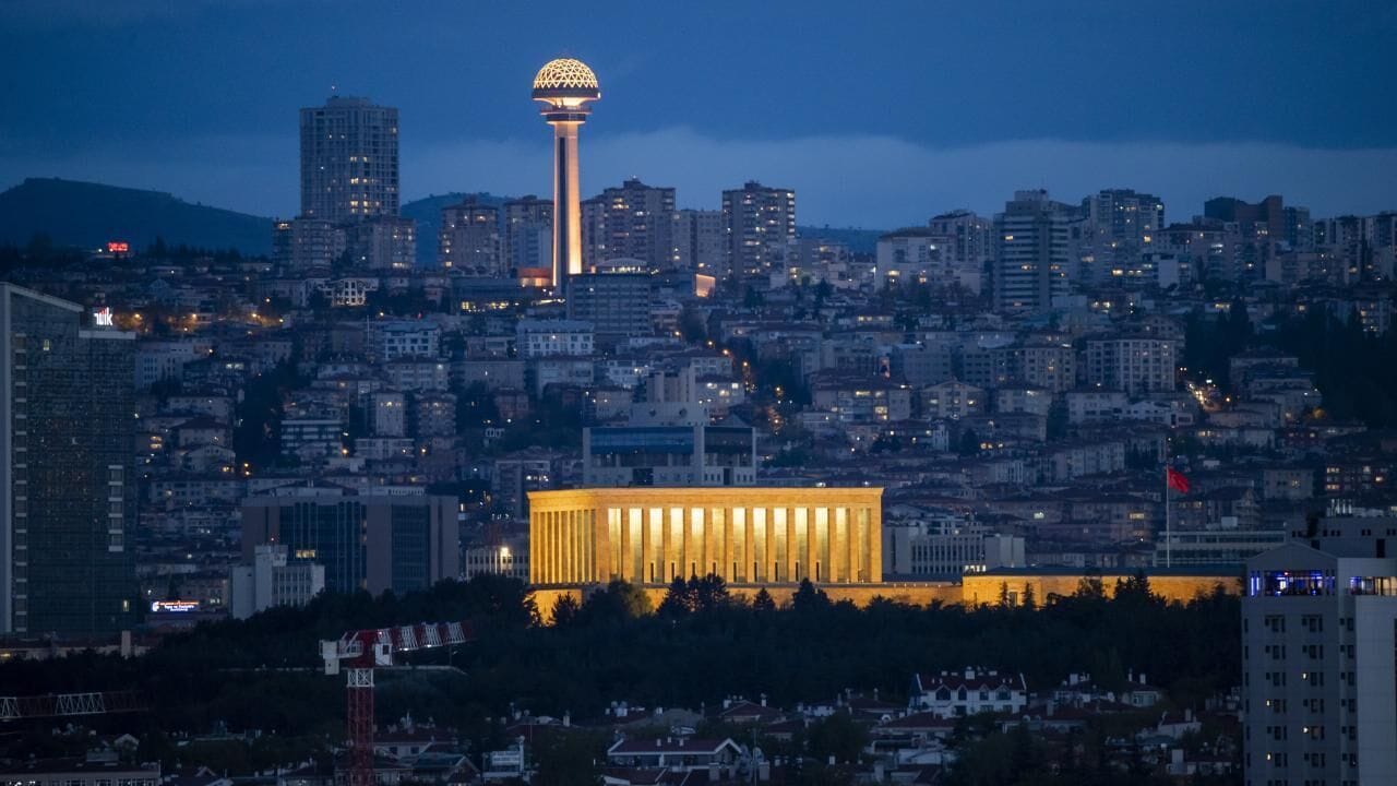 Paisagem urbana ao entardecer com um edifício imponente de linhas verticais e uma estrutura esférica ao fundo.