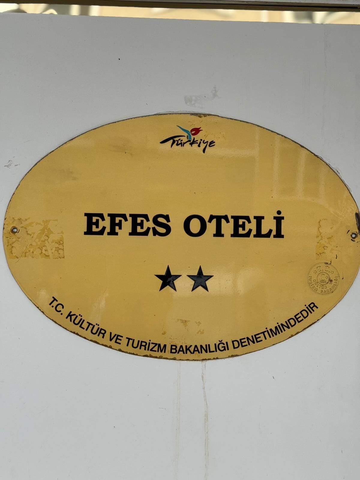 Un letrero amarillo con la inscripción 'EFES OTELI' y 'T.C. KULTUR VE TURIZM BAKANLIĞI DENETİMİNDİR', que indica un establecimiento relacionado con la cultura y el turismo.