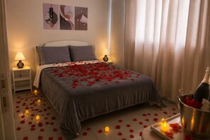 En pænt redt seng med rosenblade og stearinlys, et sengebord med en lampe, og en flaske champagne på gulvet.