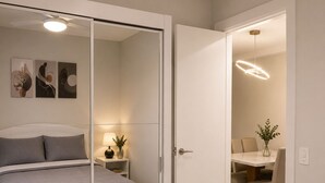 Een slaapkamer met een bed, nachtkastje met lamp, spiegel en een deur naar een eetruimte met tafel en stoelen.