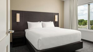 A hotel room with a large bed, a nightstand, a wall-mounted lamp, and a window with a view of trees.