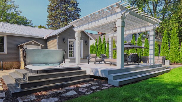 A covered outdoor patio with a hot tub, seating area, and steps leading to a house.