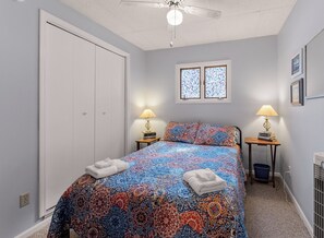 Une chambre à coucher comprenant un lit, deux tables de chevet, un ventilateur de plafond, une fenêtre et une porte.