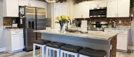 Cocina moderna con armarios blancos, refrigerador de acero inoxidable, una isla grande con taburetes y luminarias colgantes.