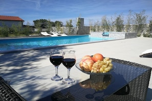 A poolside table with two glasses of red wine, a bowl of fruit, and a view of a swimming pool. - Pool Apartment Daisy 4, Privlaka, Croatia (Privlaka)