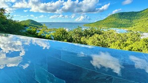Klarer, blauer Himmel, ein Pool mit Glasboden und Blick auf eine grüne Landschaft mit Hügeln und einem Gewässer.