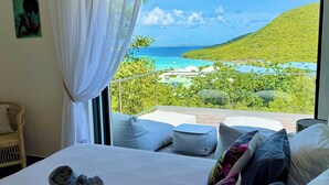 Ein Schlafzimmer mit großem Fenster, einem Bett mit Kissen, einem Sessel und Blick auf eine Landschaft.