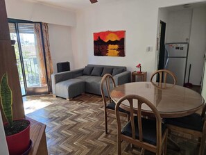 A living room with a herringbone floor, a grey sectional sofa, a round wooden dining table with chairs, and a painting of a sunset. - Apartment. 2 bdrm. w / balcony V. Crespo (Villa Crespo)
