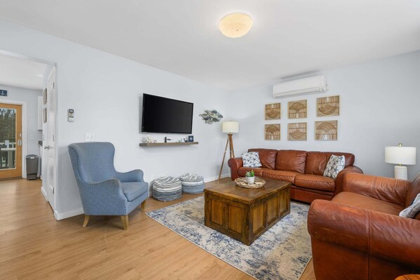 Ein Wohnzimmer mit blauem Sessel, braunem Ledersofa, Holztisch, Wandfernseher und Wandkunst.