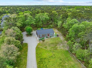 A house surrounded by dense forest, with a gravel driveway leading to the front door. - Charming Laid-Back Cape Escape (Harwich)