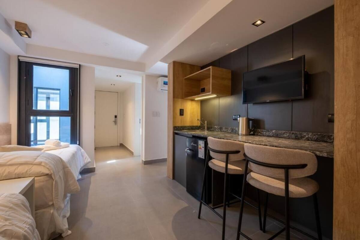 Een moderne hotelkamer met een kitchenette, een aan de muur bevestigde televisie en een groot raam.