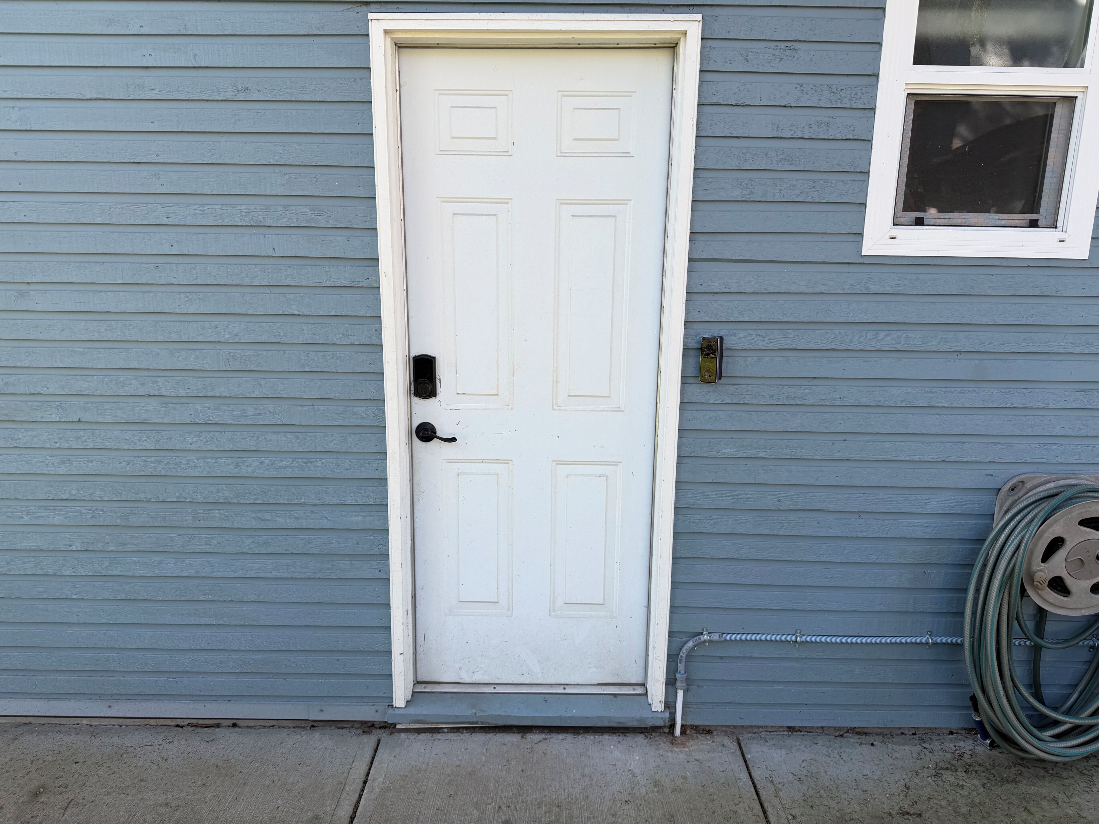 A white door with a black handle, a window with a metal grill, and a coiled garden hose against a blue siding house.