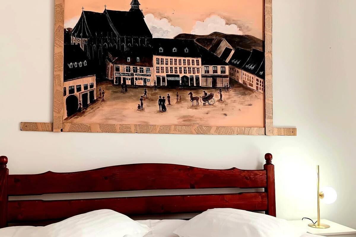 A hotel room with a bed, pillows, and a painting of a town square.
