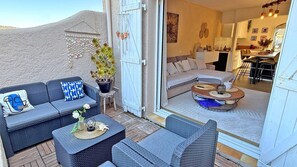Un rincón exterior acogedor con mobiliario de mimbre, una planta en maceta y vistas hacia un interior moderno con sofá y comedor.
