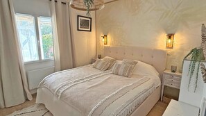 Un dormitorio con una cama grande, dos mesitas de noche, una lámpara colgante y una ventana con cortinas.