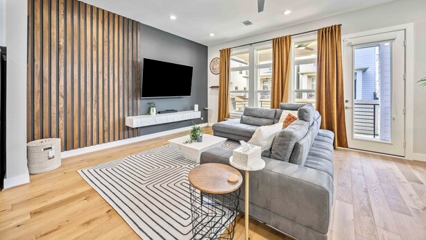 Ein modernes Wohnzimmer mit grauem Sofa, Flachbildfernseher, einer Wandpaneele aus Holz und einem Deckenventilator.