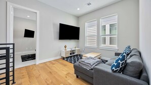 Ein modernes Wohnzimmer mit grauem Sofa, einem Flachbildfernseher, einem kleinen Holztisch und einem Fenster mit weißen Fensterläden.
