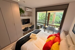 A modern bedroom with a large bed, a TV, a desk, and a balcony with outdoor furniture. - Dommy (São Paulo)