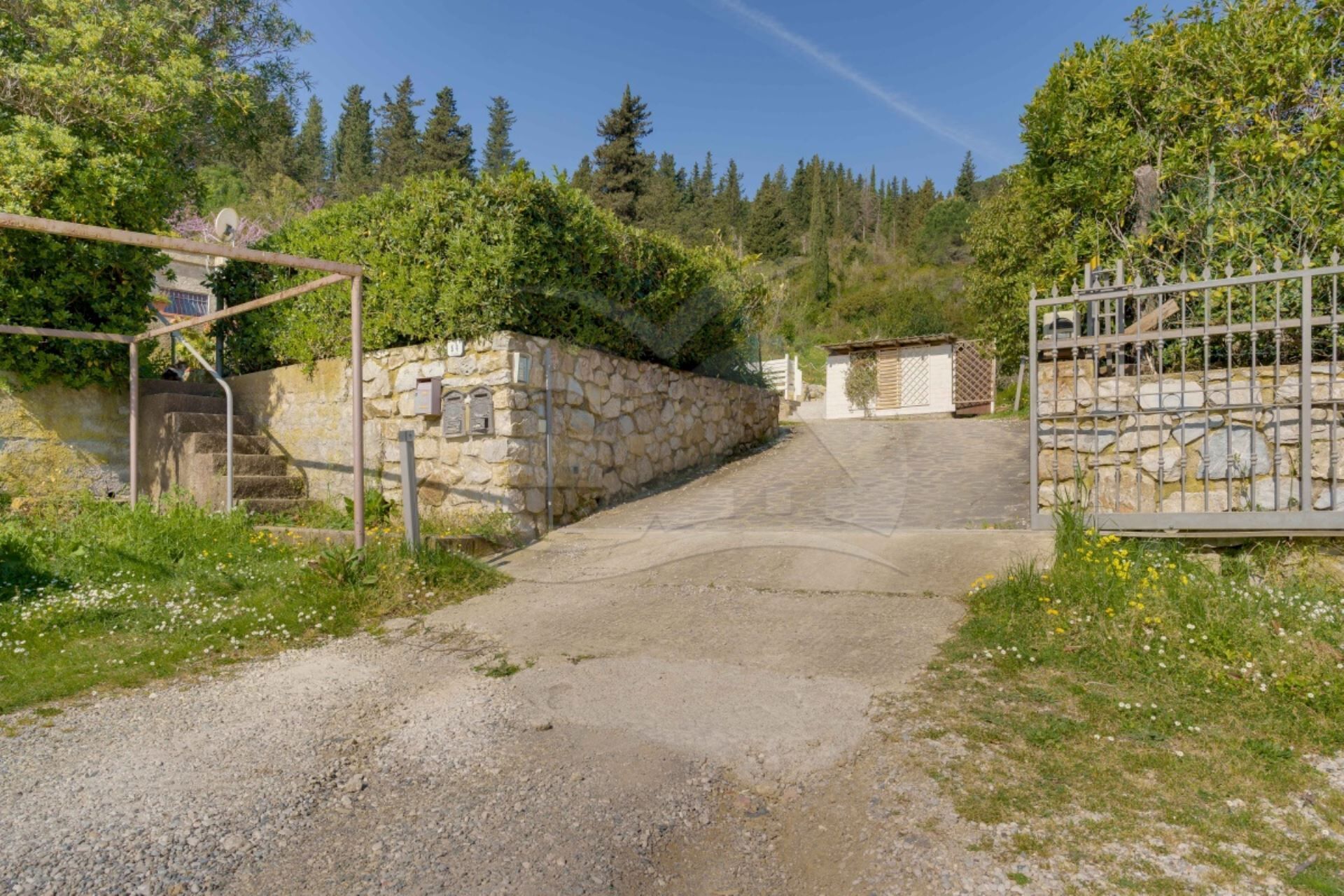 Une allée de gravier menant à un mur de pierres surmonté d’un portail en métal.