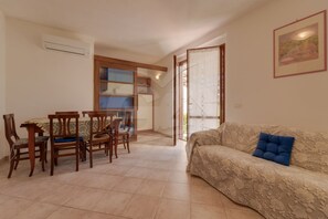 A living room with a sofa, a dining table with chairs, a kitchen area, and a door leading outside. - VILLETTA LA COLOMBAIA (Porto Azzurro)