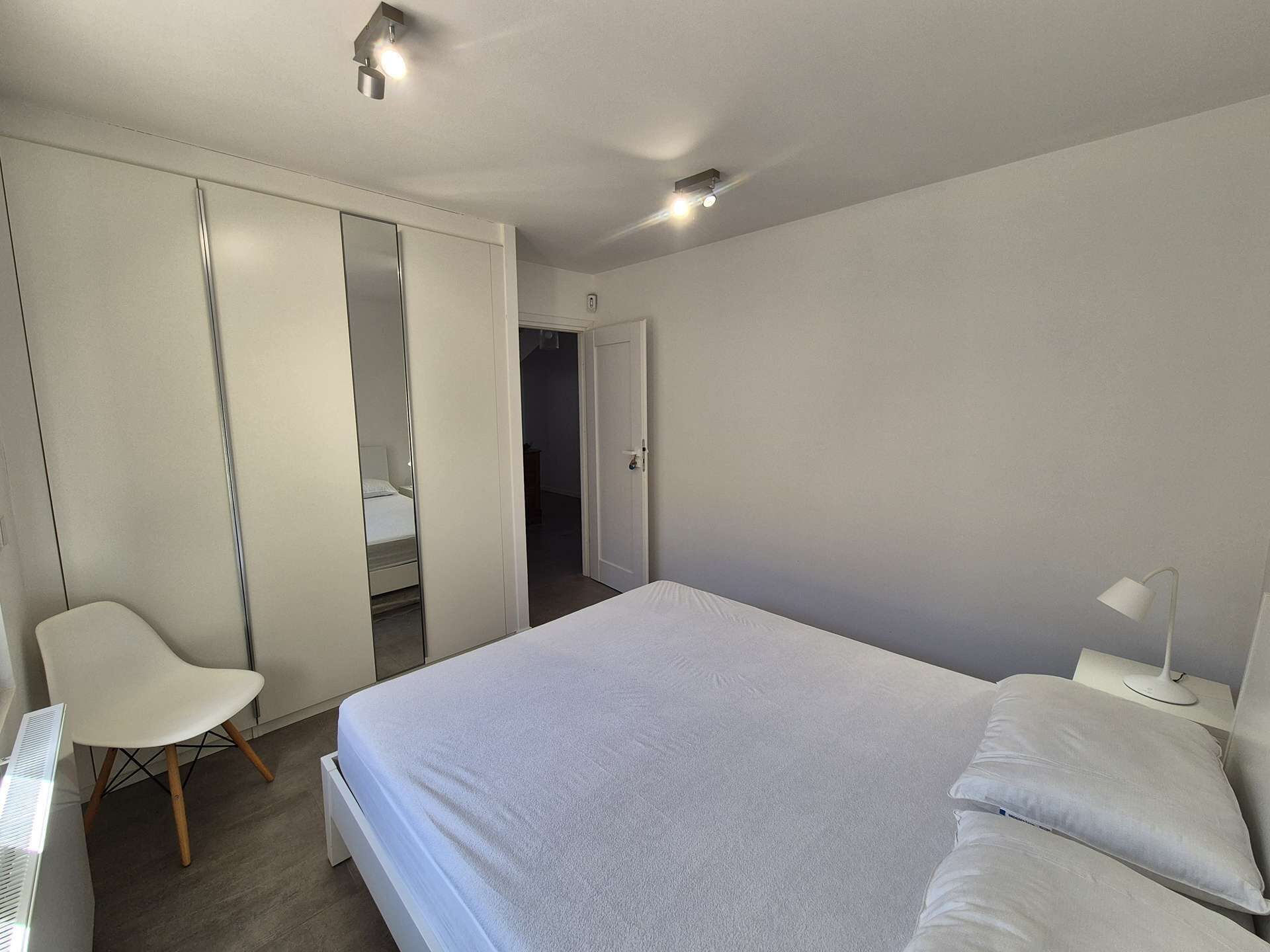 A bedroom with a bed, a chair, a wardrobe, and a nightstand with a lamp.