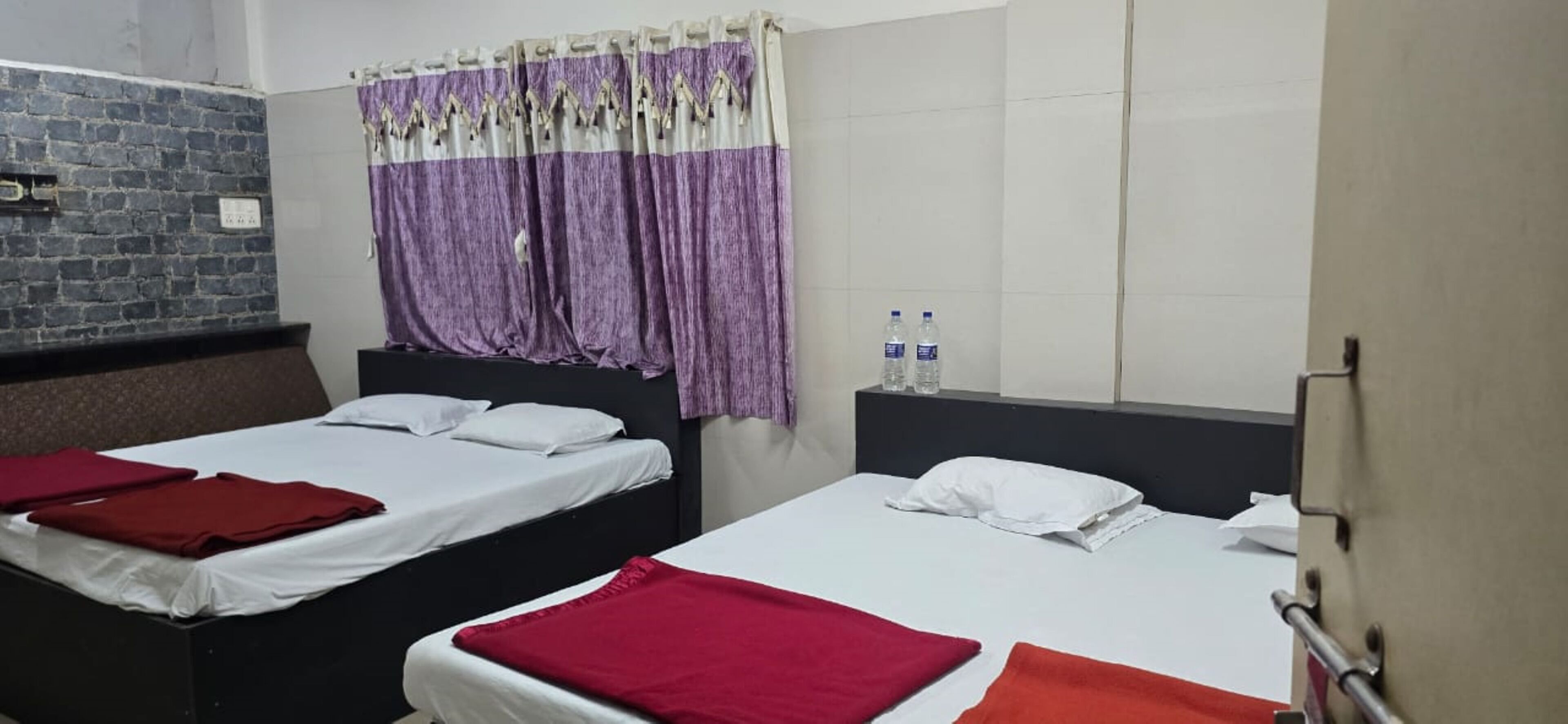 A hotel room with two beds, a brick wall, a window with purple curtains, and two water bottles on the nightstand.