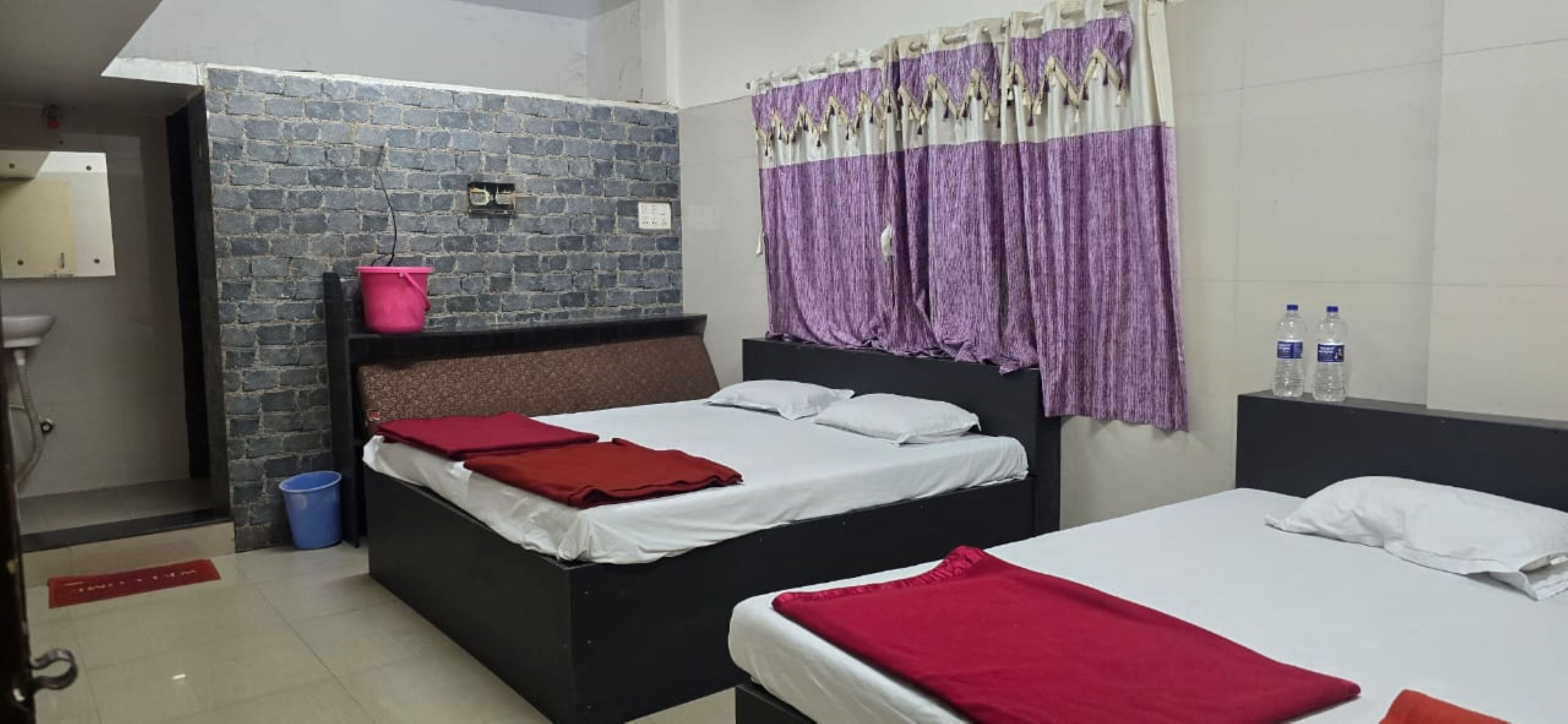 A hotel room with two beds, a brick accent wall, a purple curtain, and a red mat on the floor.