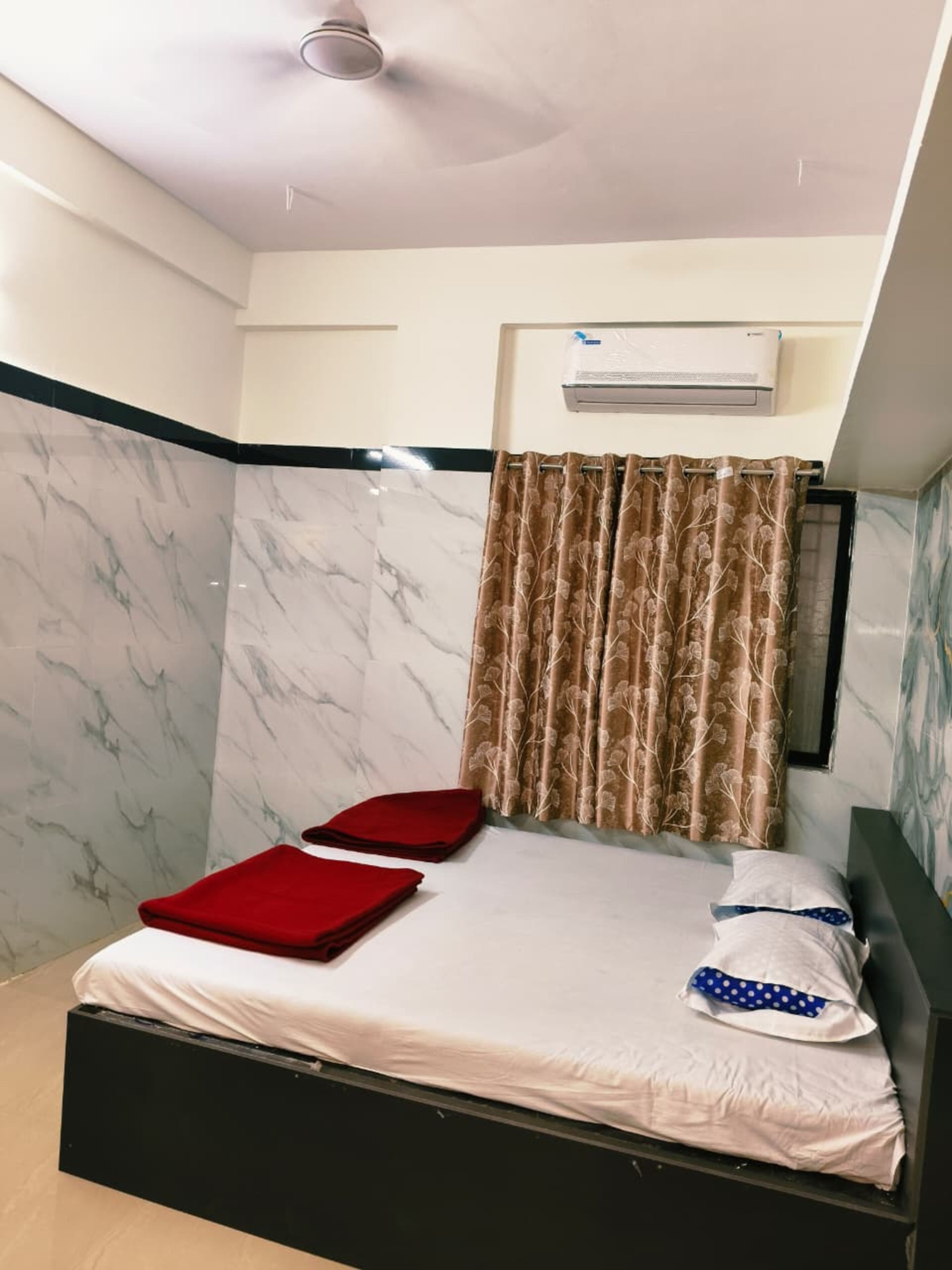 A single bed with a white mattress, red pillows, and a blue polka-dotted pillow. A brown floral curtain is drawn across the window.