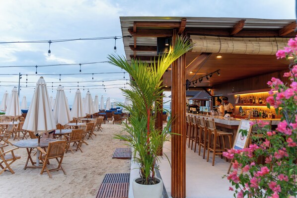 An outdoor seating area with wooden tables and chairs, umbrellas, and a bar area with a counter and stools.
