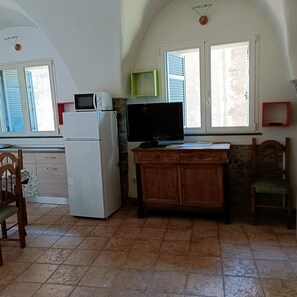 Eine kleine Küche mit Kühlschrank, Mikrowelle und einem Flachbildfernseher. Es gibt Fenster mit Rollläden, einen Holzkasten und einen Stuhl.