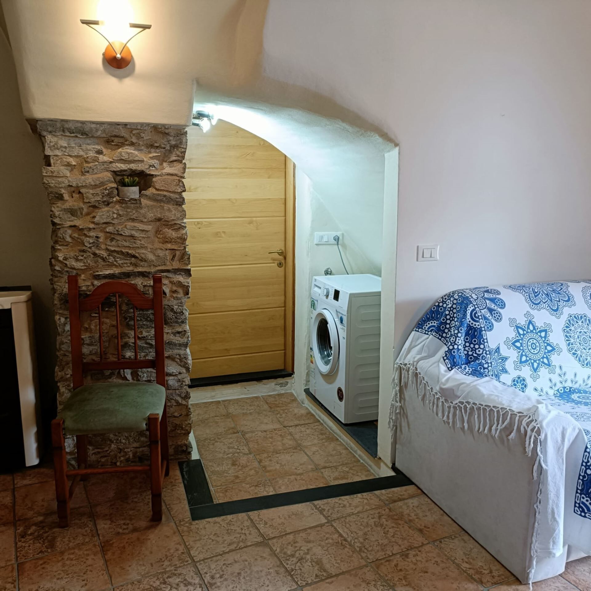 A room with a stone wall, wooden door, washing machine, bed with patterned bedding, and a chair.