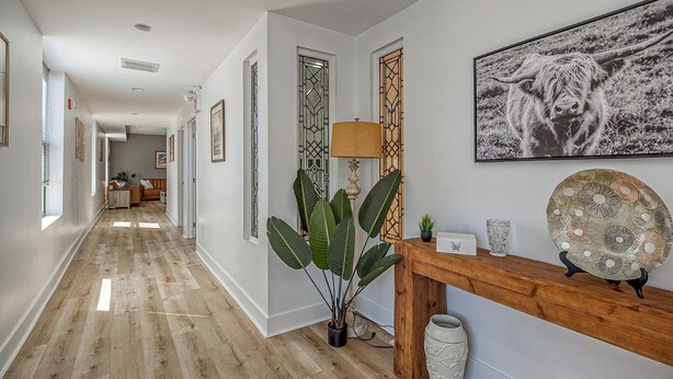 A hallway with a wooden floor, a potted plant, a lamp, a decorative mirror, and a framed picture on the wall. - Cozy Carnegie Retreat • Minutes to Downtown PGH (Carnegie)