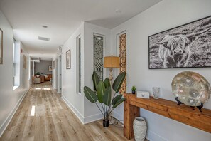A hallway with a wooden floor, a potted plant, a lamp, a decorative mirror, and a framed picture on the wall. - Cozy Carnegie Retreat • Minutes to Downtown PGH (Carnegie)