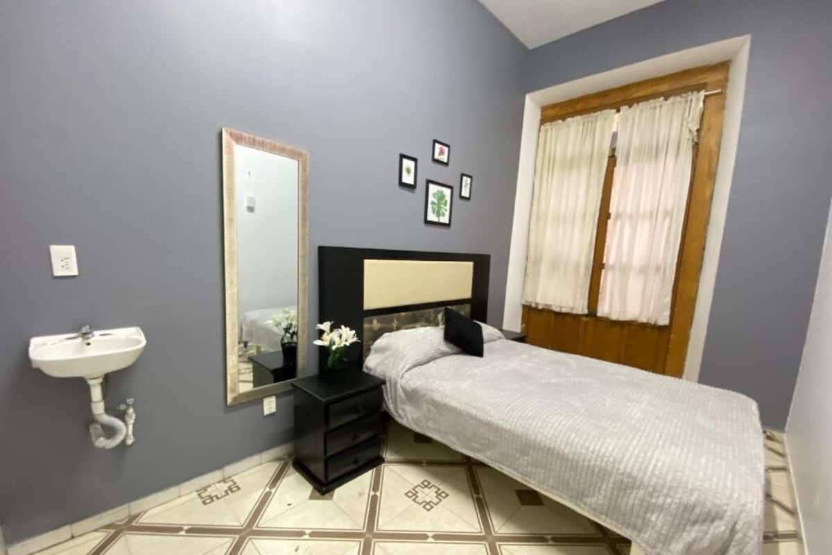A bedroom with a bed, a bedside table, a mirror, a window with curtains, a sink, and a wall with framed pictures.