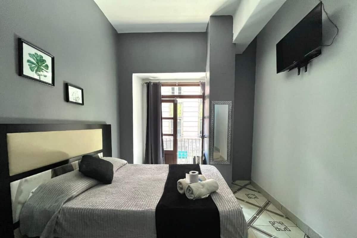 A hotel room with a bed, a TV, a mirror, and a view of the outside.
