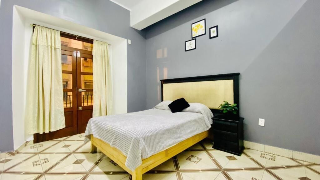 A bedroom with a bed, a nightstand, a window with curtains, and a wall with framed pictures.