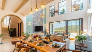 A spacious dining area with a wooden table set for a meal, a television mounted on the wall, and large windows offering a view of the outdoors.