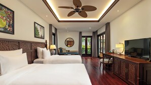 Quarto de hotel com cama, televisão, escrivaninha com luminária, cadeira, sofá, ventoinha no teto e uma janela grande.