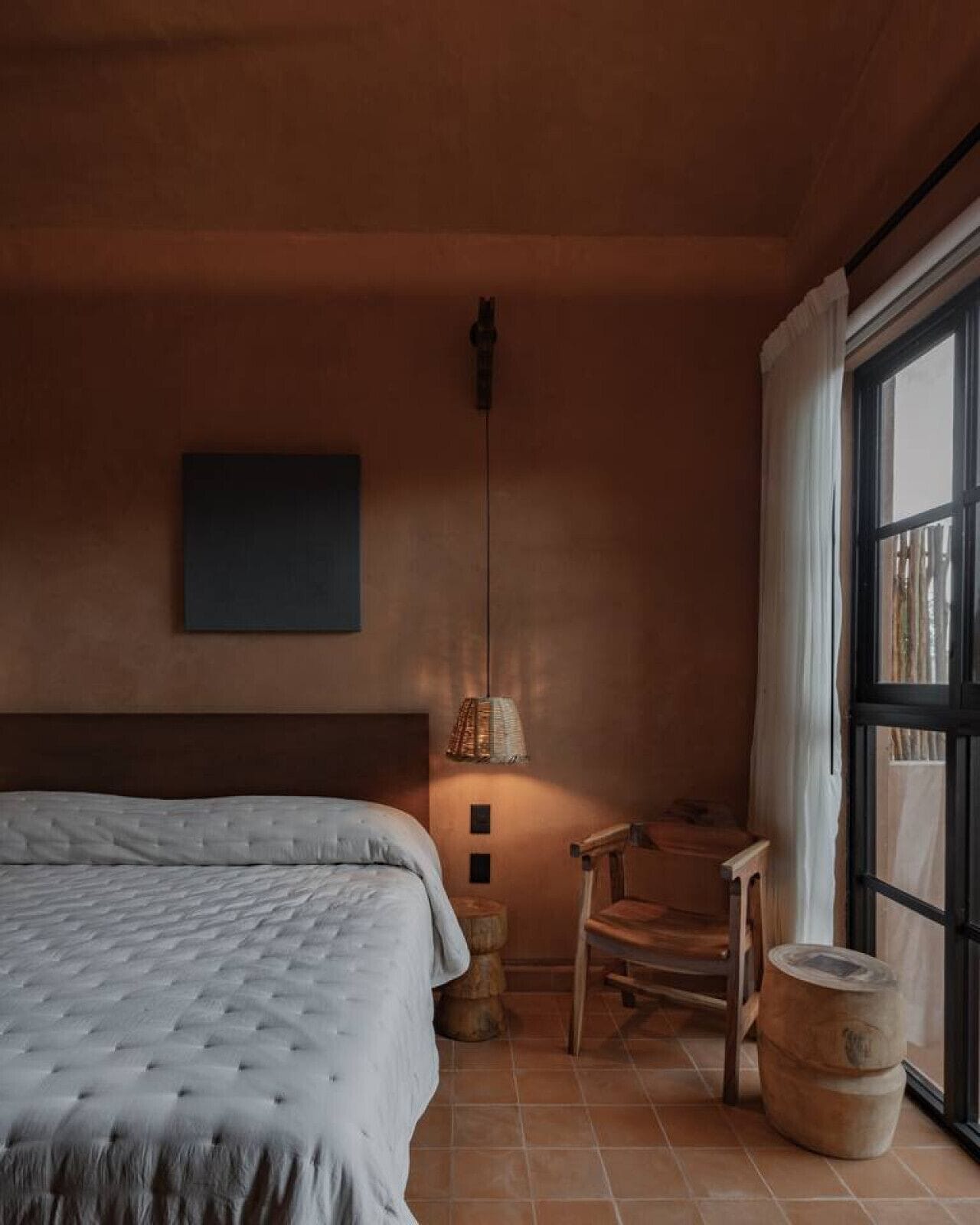 A bedroom with a bed, a chair, a table, a lamp, and a large window.