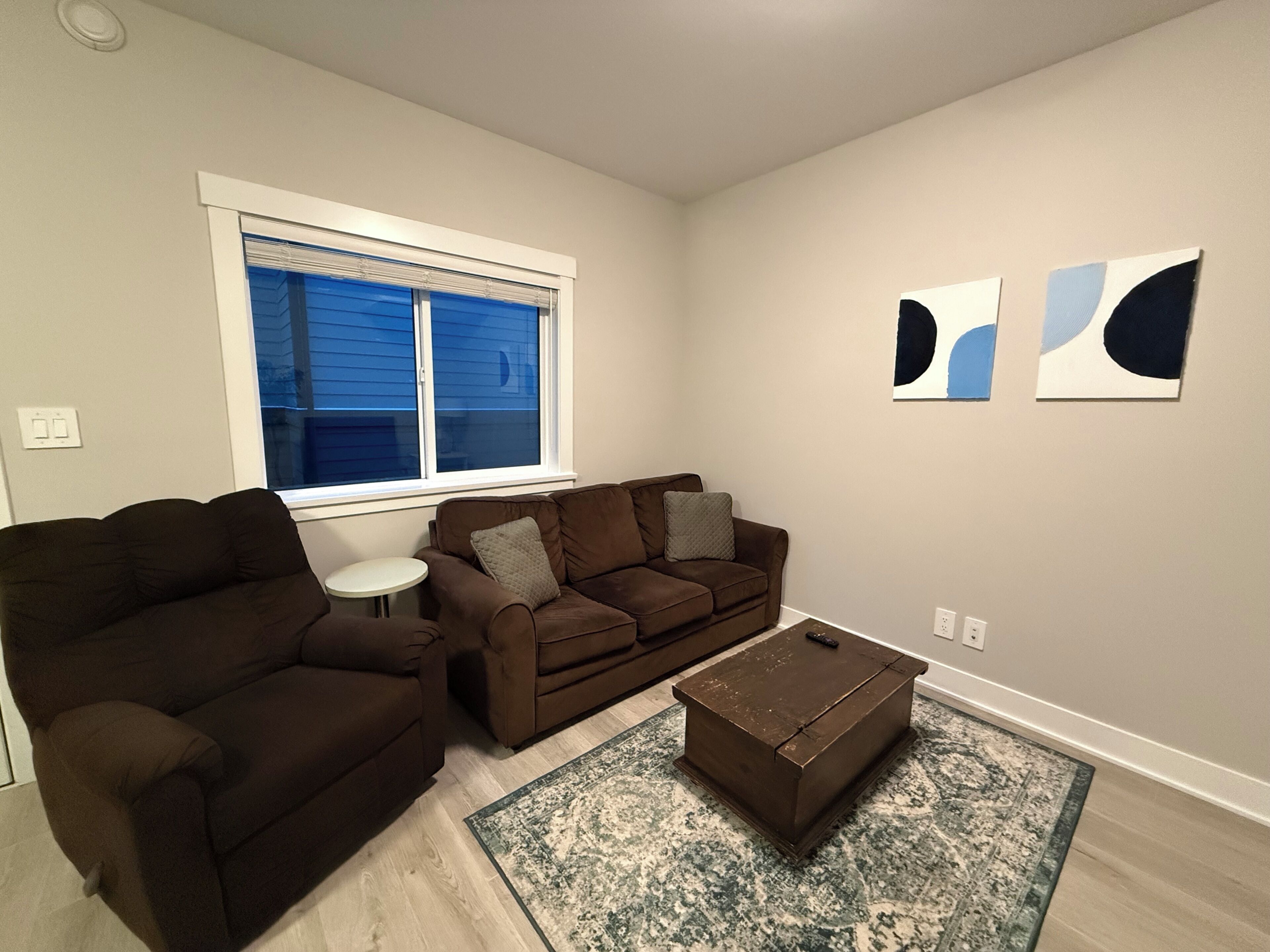A living room with a brown sofa, a small white table, a coffee table, and abstract wall art.