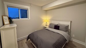 Un dormitorio con cama, mesita de noche, lámpara, ventana y estante en la pared con un marco de fotos.