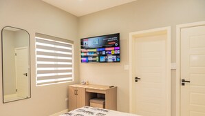 A bedroom with a bed, a TV mounted on the wall, a mirror, and a window with blinds. - Blue Haven Suite at Black Pineapple Cottages (Cedar Grove)