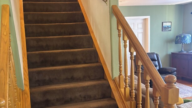 A carpeted staircase with wooden balusters, a door at the top, and a room with a chair and lamp.