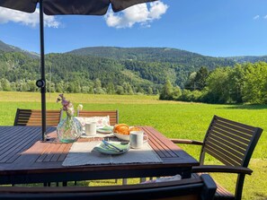 Uma mesa de piquenique com um vaso, pratos e um croissant, com um cenário de um campo verde e colinas arborizadas.