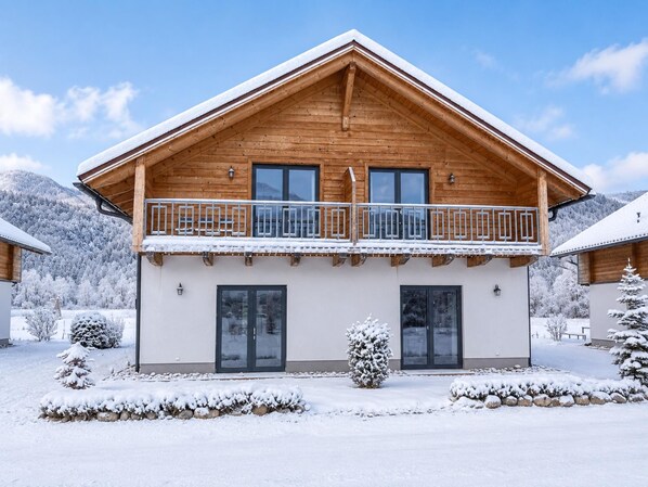 Un chalet de madera de dos pisos con balcón, ventanas grandes y un paisaje cubierto de nieve.