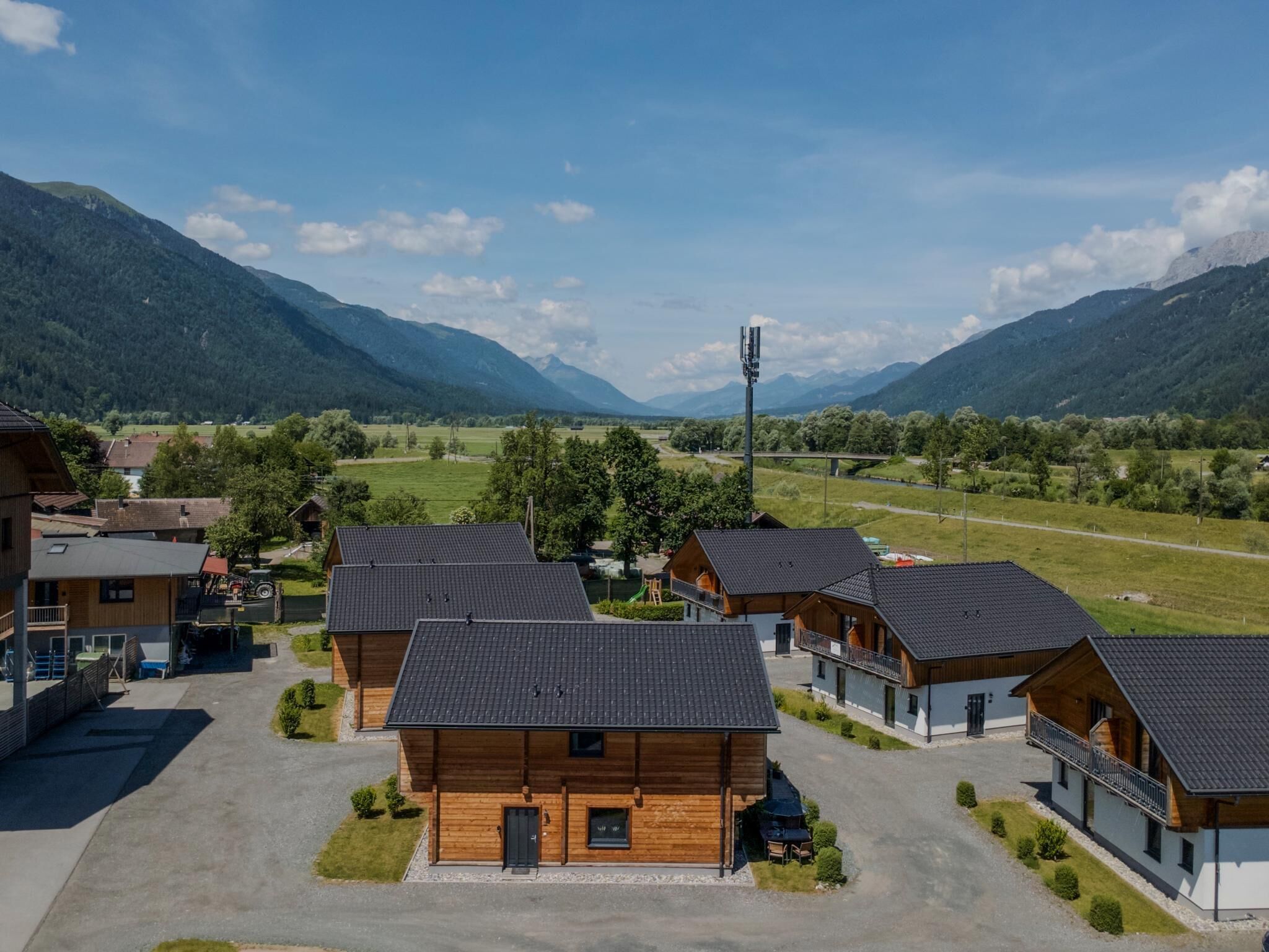 A cluster of wooden cabins with a paved driveway, surrounded by lush greenery and mountains in the background.