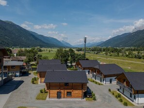 Un ensemble de chalets en bois, avec une allée pavée, entouré d’une végétation luxuriante et bordé de montagnes en arrière-plan.